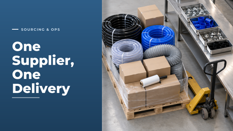 High-angle view of a hose distributor despatch floor with one mixed pallet of black rubber, blue silicone and clear braided PVC hose coils, grey ducting and cartons being stretch-wrapped, beside a packing bench of hose clips and silicone joiners