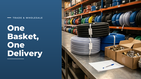 One consolidated trade order grouped on a stainless steel counter in a hose distribution warehouse: banded coils of black rubber, clear braided PVC and blue layflat hose beside open boxes of hose clips and couplings, with racked hose behind
